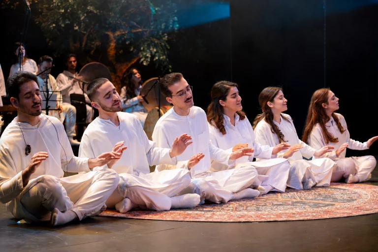 Zendeh Delan members in white seated in a row on a carpet during the Zekr (dhikr) scene of The Conference of the Birds in Dallas